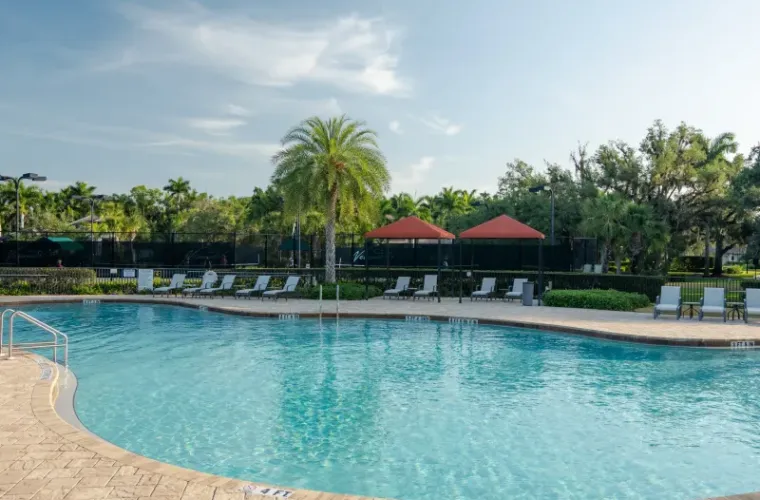 A large freeform community pool surrounded by lounge chairs, palm trees, and a tennis court fence under a sunny sky.