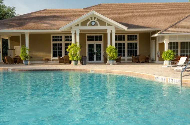 Community clubhouse pool deck with lounge chairs and potted plants in front of a Spanish-tile-roofed building.