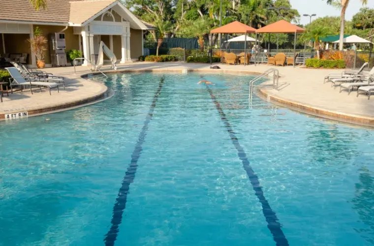 A lap pool with lane markers at a community clubhouse with a distant swimmer, surrounded by lounge chairs and umbrellas.