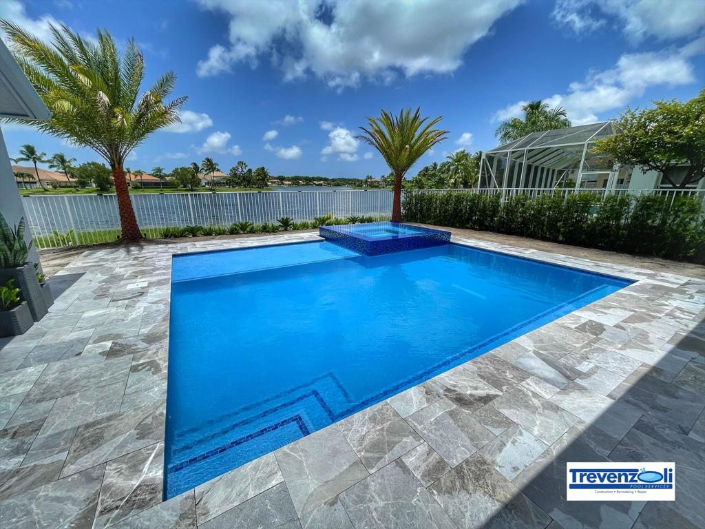 A rectangular backyard pool with a raised tiled spa overlooking a lake, framed by palm trees and stone pavers.